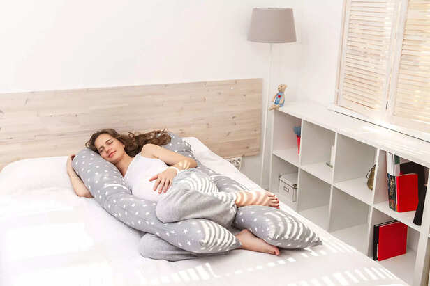 Beautiful sleeping smiling pregnant woman with a big tummy abdomen with long curly hair and closed eyes, hugging a long body pillow with her arm and legs and lying on the bed indoor.